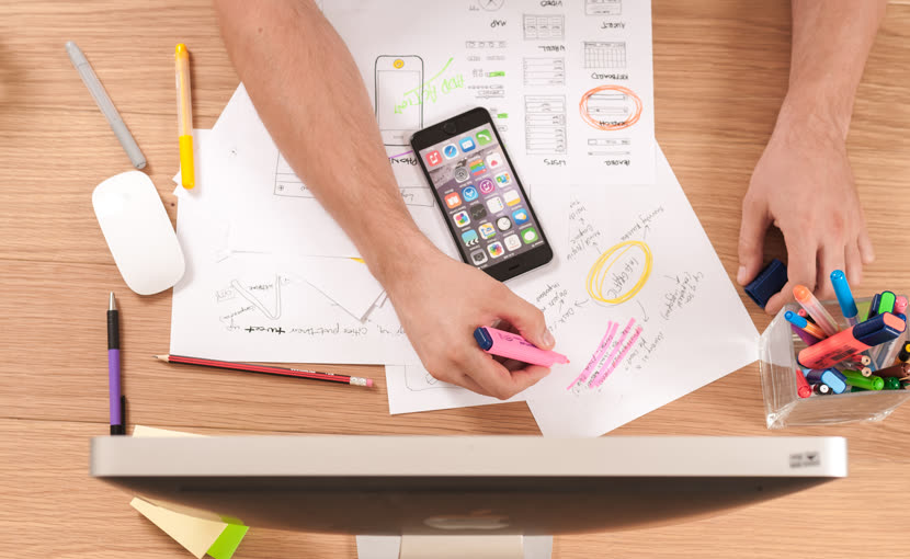 Hands writing at a desk beside a phone, pens and a monitor
